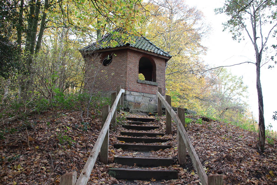 Bakstenen tuinhuis met groen dak, bereikbaar via trap, omgeven door herfstbomen en bladeren.