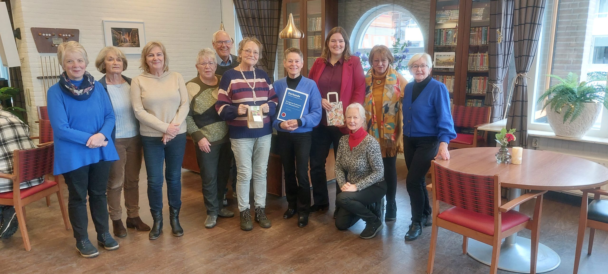 Een groep mensen poseert in een kamer met boekenplanken en een tafel met bloemen.