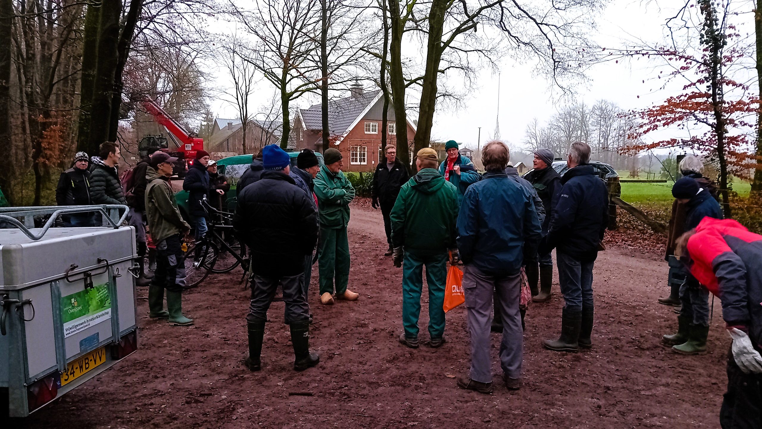 Groep mensen in regenjassen verzamelt zich op een modderig bospad bij een tractor en een huis.