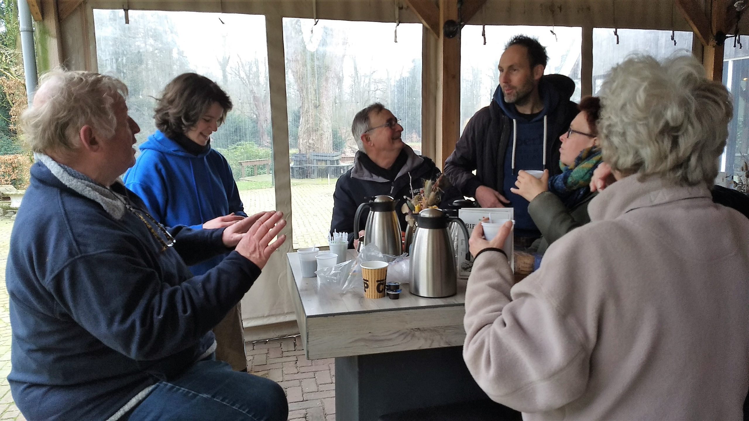 Groep mensen zit rondom een tafel met thermoskannen, pratend en lachend in een overdekt buitenruimte.
