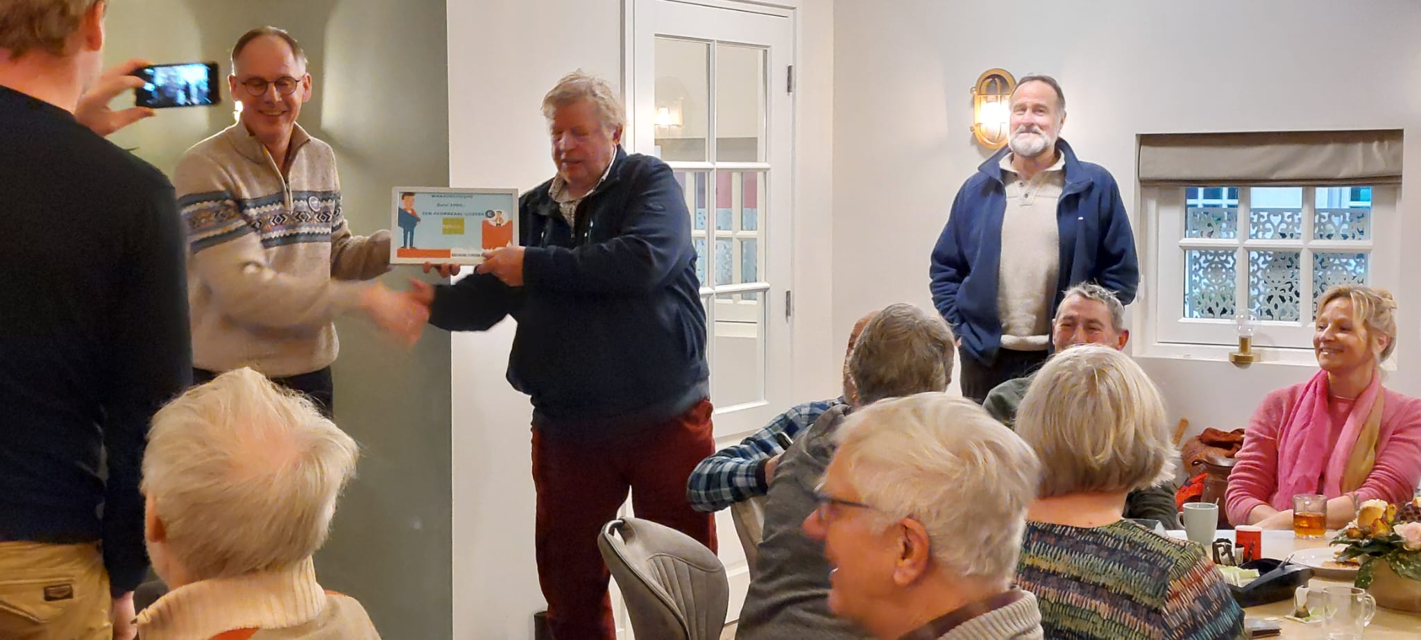 Man ontvangt certificaat in volle zaal; mensen kijken glimlachend toe.