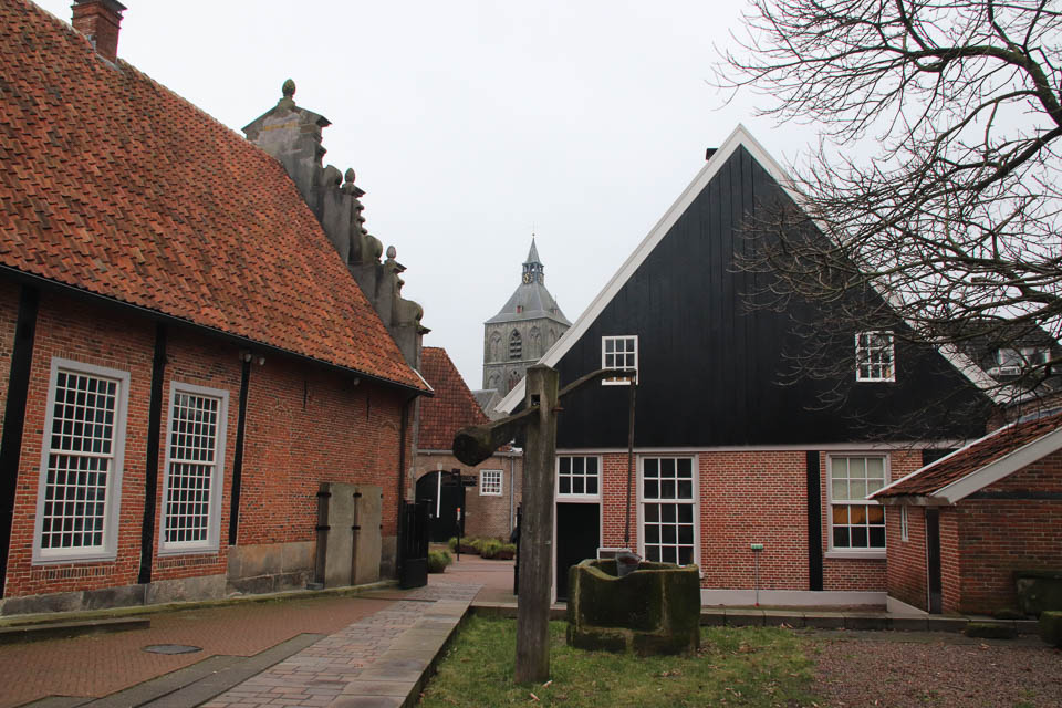 Historisch gebouw met rode bakstenen, een zwart zadeldak en een torenspits op de achtergrond.