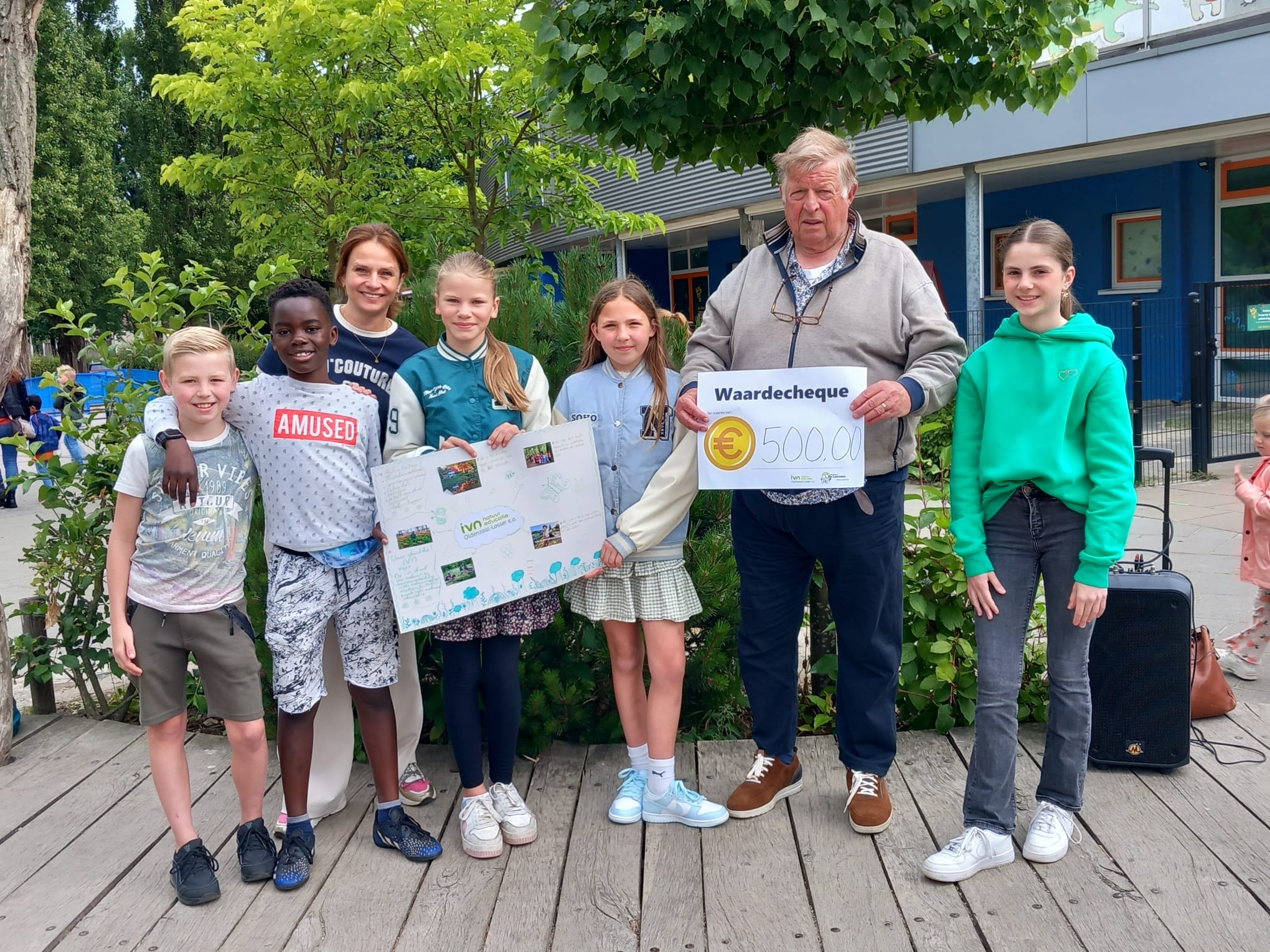 Groep kinderen en volwassene met cheque van 500 euro, buiten bij een schoolgebouw.