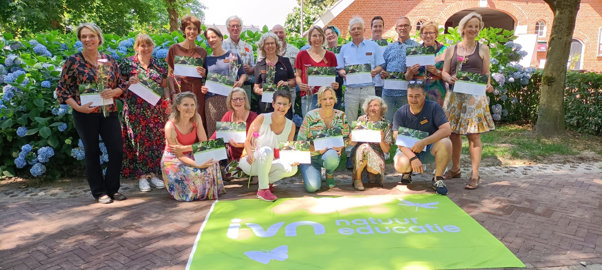 Groep mensen met certificaten en bloemen, poseren voor een IVN natuureducatie banner buiten.