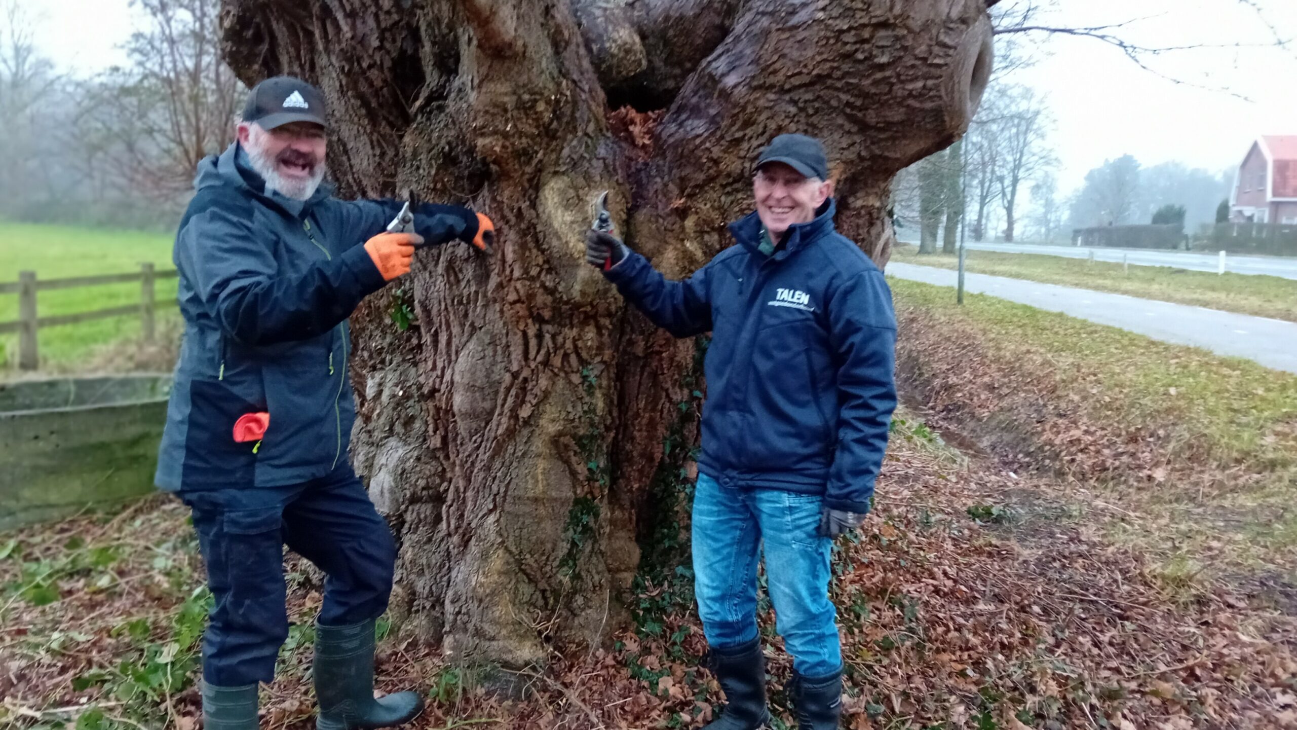 Twee mannen naast een dikke boom, beiden dragen handschoenen en werkjassen, lachend naar de camera.
