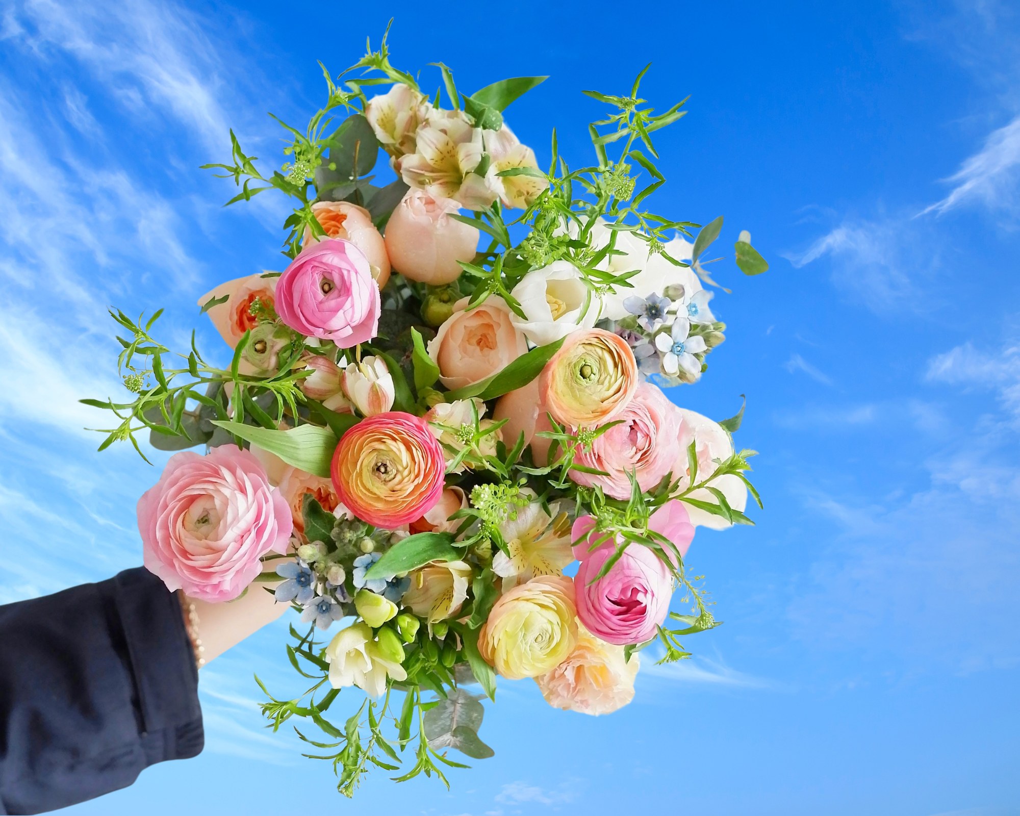Hand houdt bloemenboeket tegen blauwe lucht met wolken.