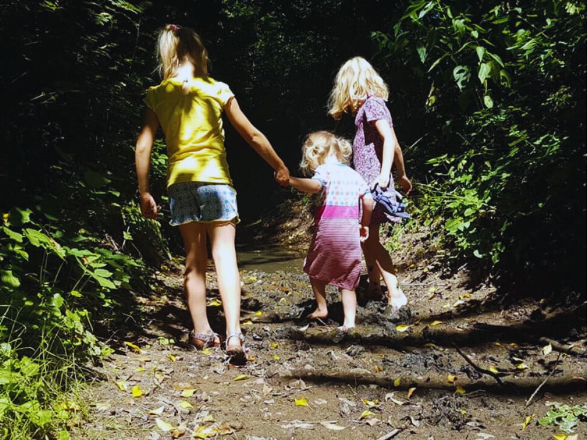 Drie kinderen lopen hand in hand op een modderig bospad, omringd door groen.