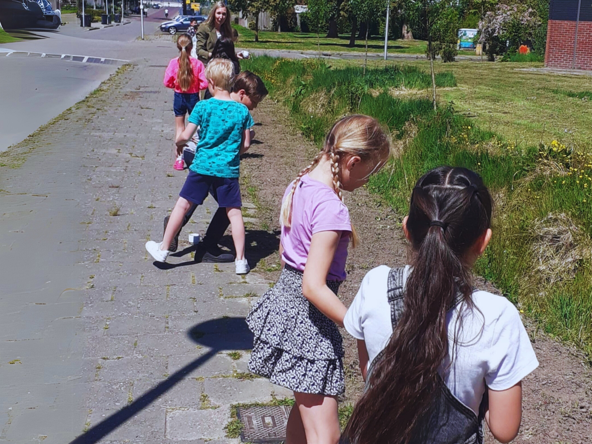 Kinderen onderzoeken planten langs een stoep met een volwassene in de buurt.