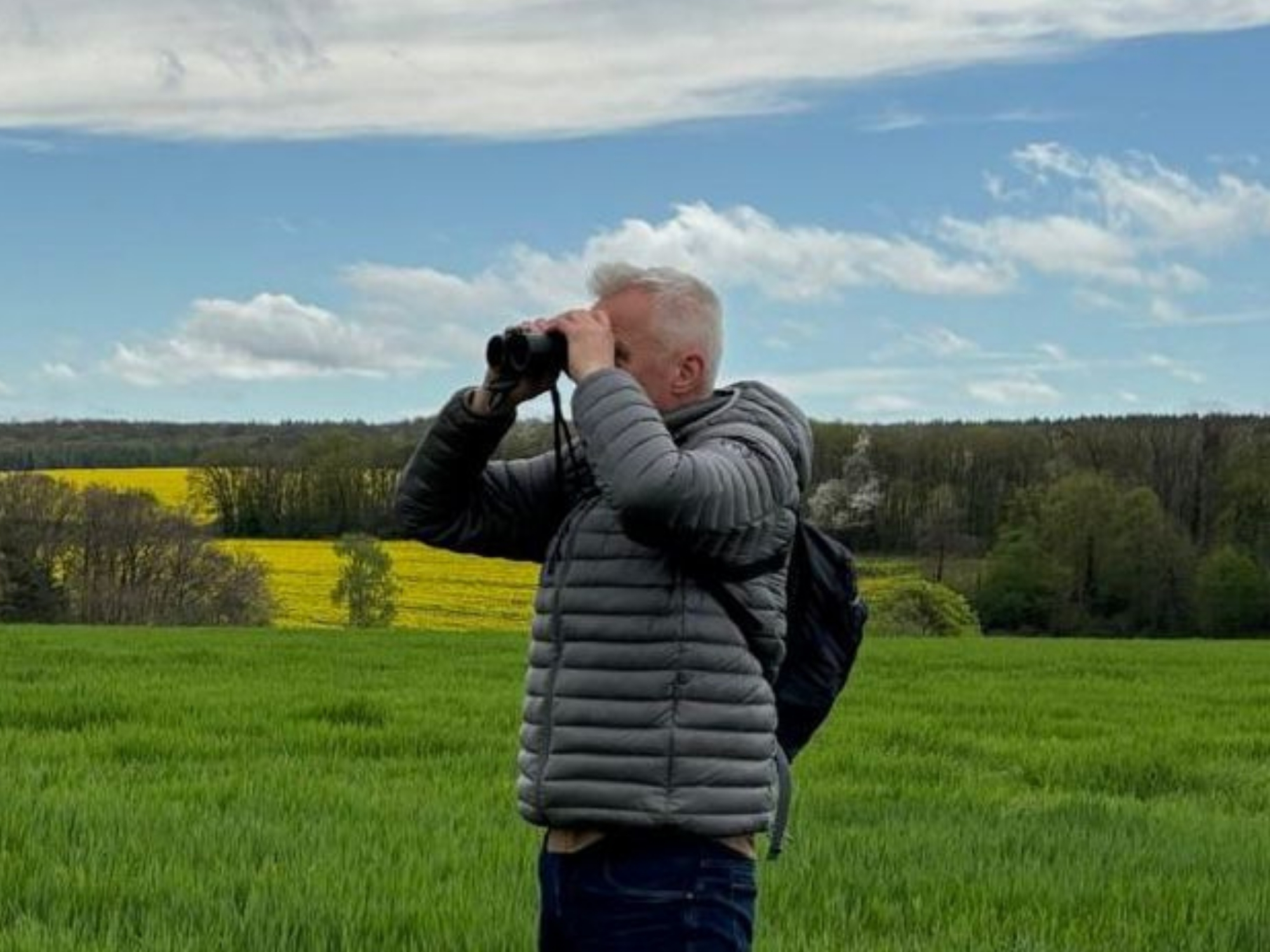 Man met verrekijker kijkt naar de lucht in een groen veld met bomen en bloemen op de achtergrond.