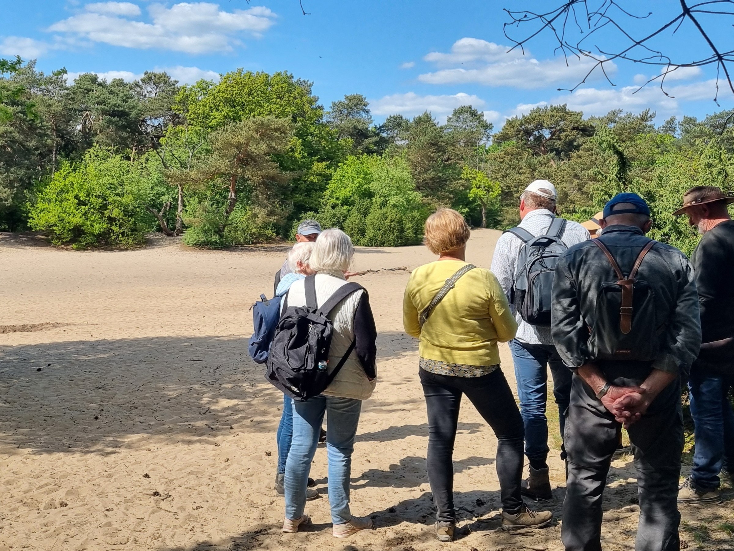 Een groep mensen staat op zandgrond bij een bosrijke omgeving onder een blauwe hemel met wolken.