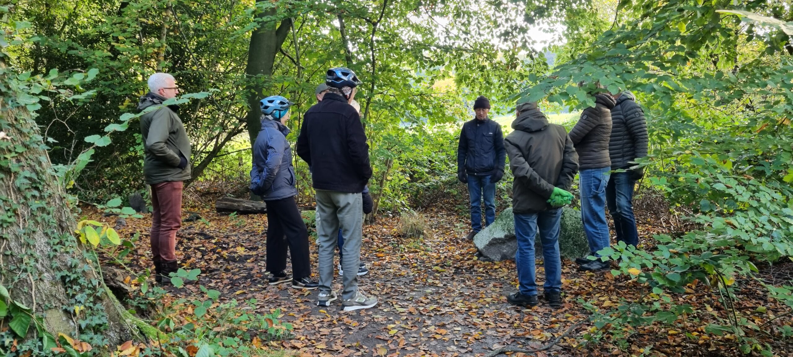 Een groep mensen in een bos, omringd door groene bladeren en herfstbladeren op de grond.