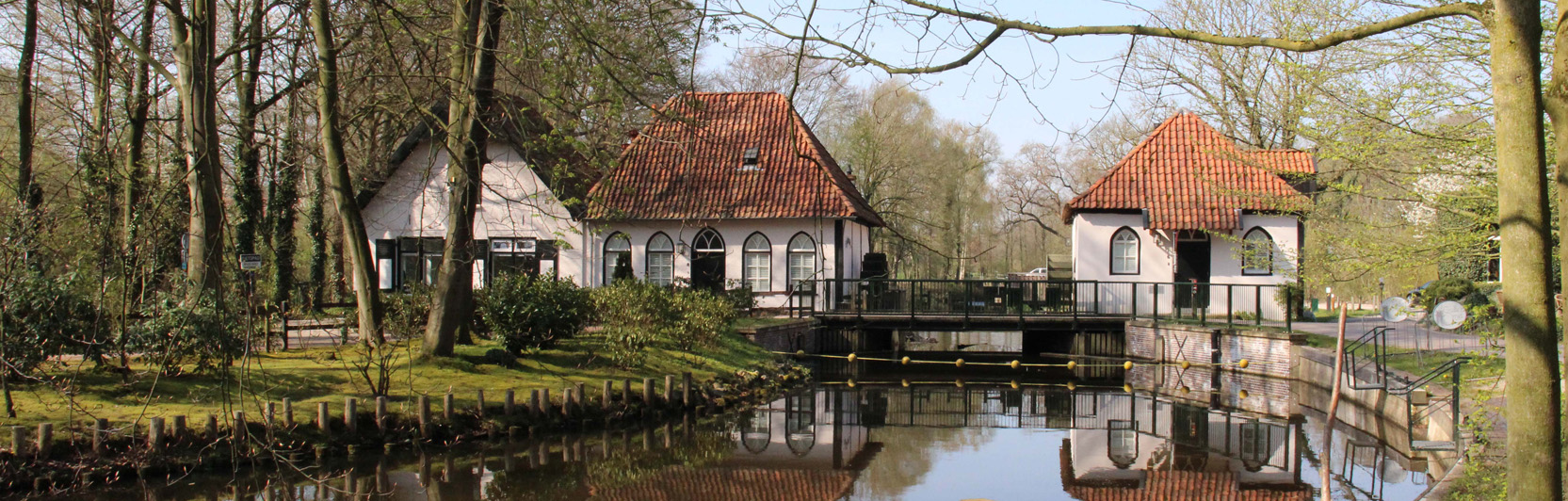 Wit huis met rode dakpannen, omringd door bomen, weerspiegeld in een vijver.