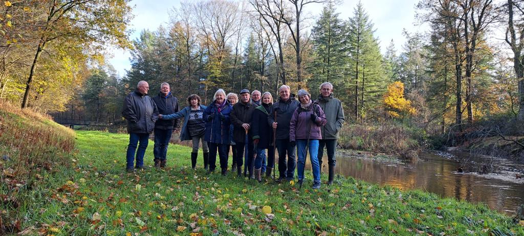 Groep senioren wandelt langs een rivier in een bosrijk herfstlandschap.
