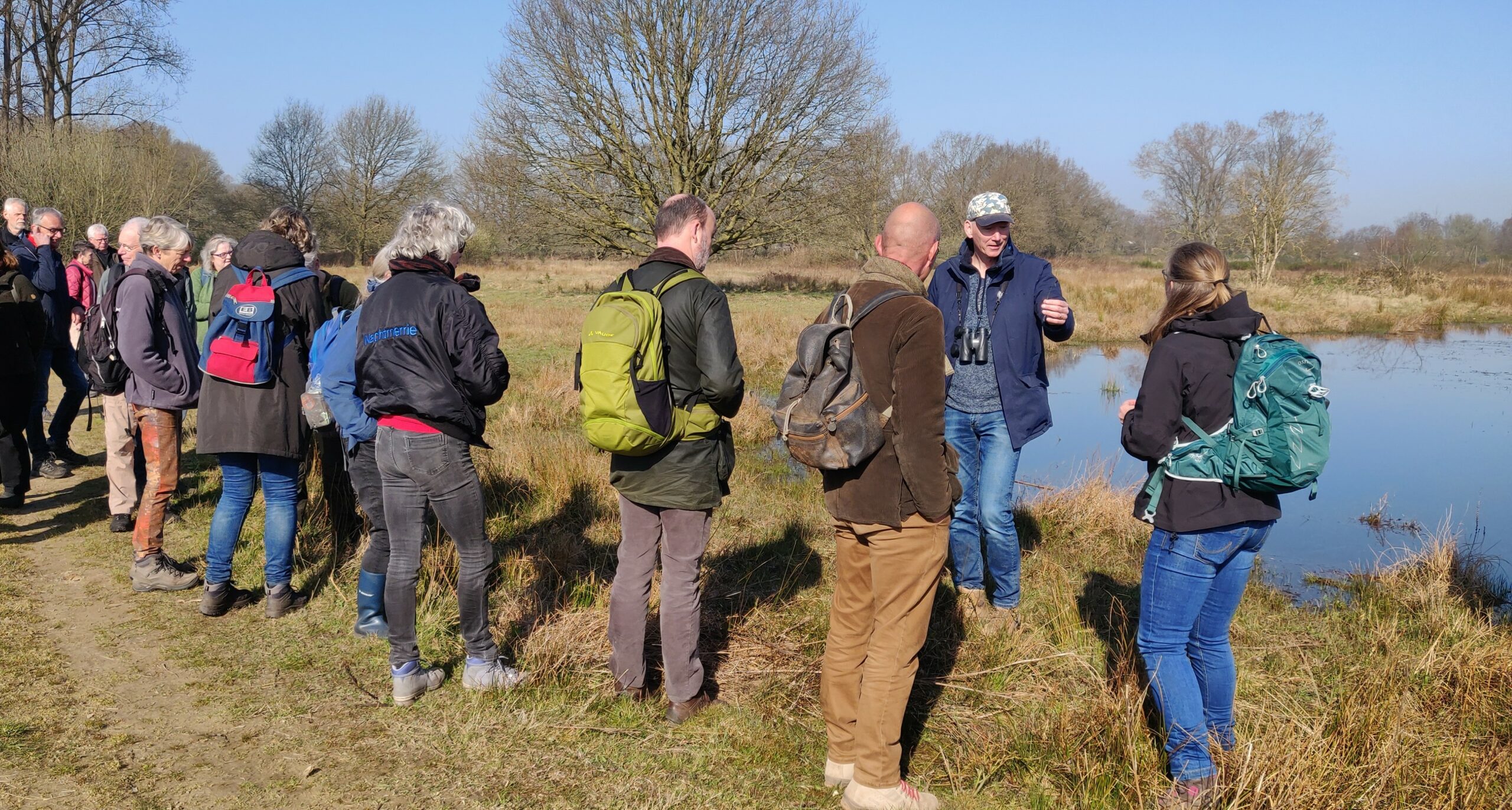 Een groep mensen met rugzakken luistert naar een gids bij een vijver in een bosrijke omgeving.