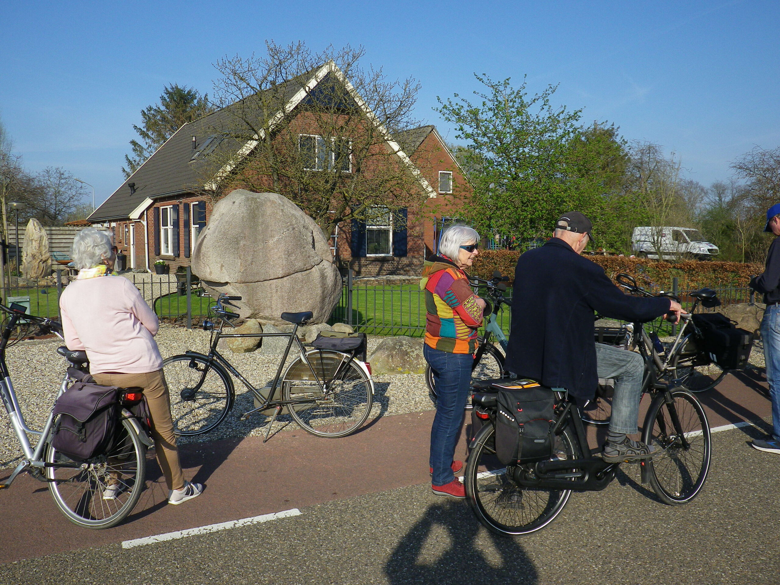 Groepje mensen op fietsen voor een huis, naast een groot rotsblok in een rustige, landelijke omgeving.