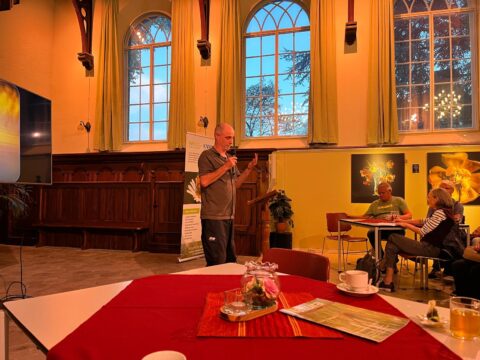 Man spreekt voor publiek in zaal met grote ramen, tafel en kunstwerken op de achtergrond.
