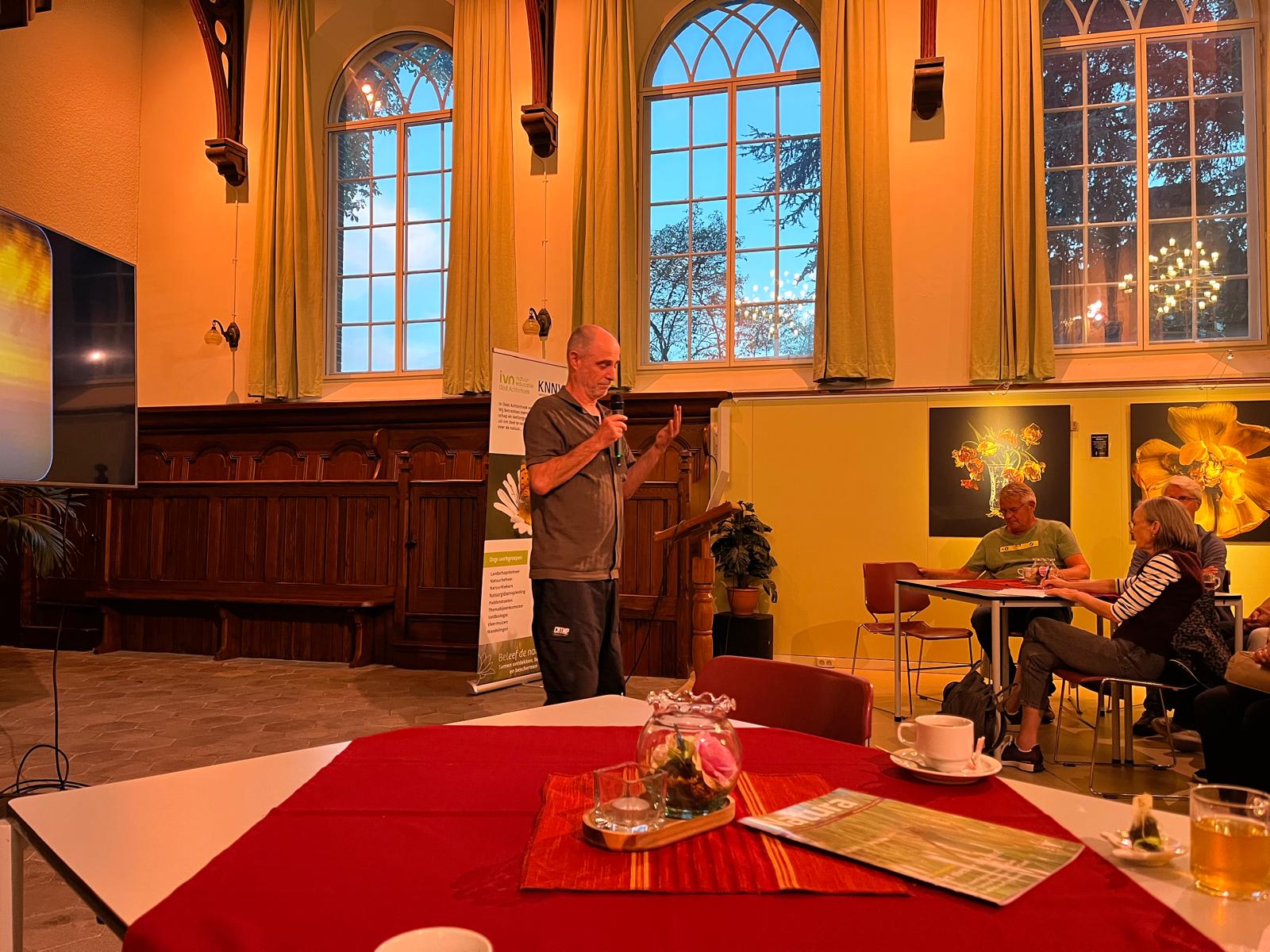 Man spreekt voor publiek in zaal met grote ramen, tafel en kunstwerken op de achtergrond.