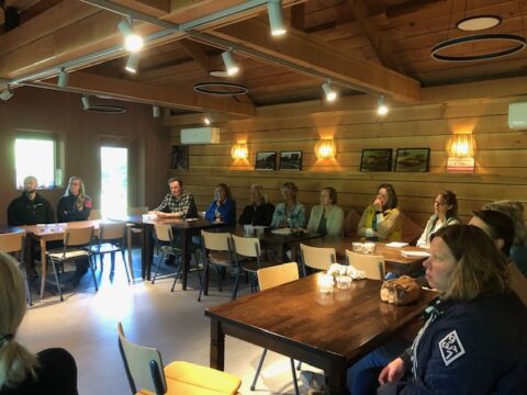 Groep mensen zit aan tafels in een houten ruimte bij een vergadering of presentatie.