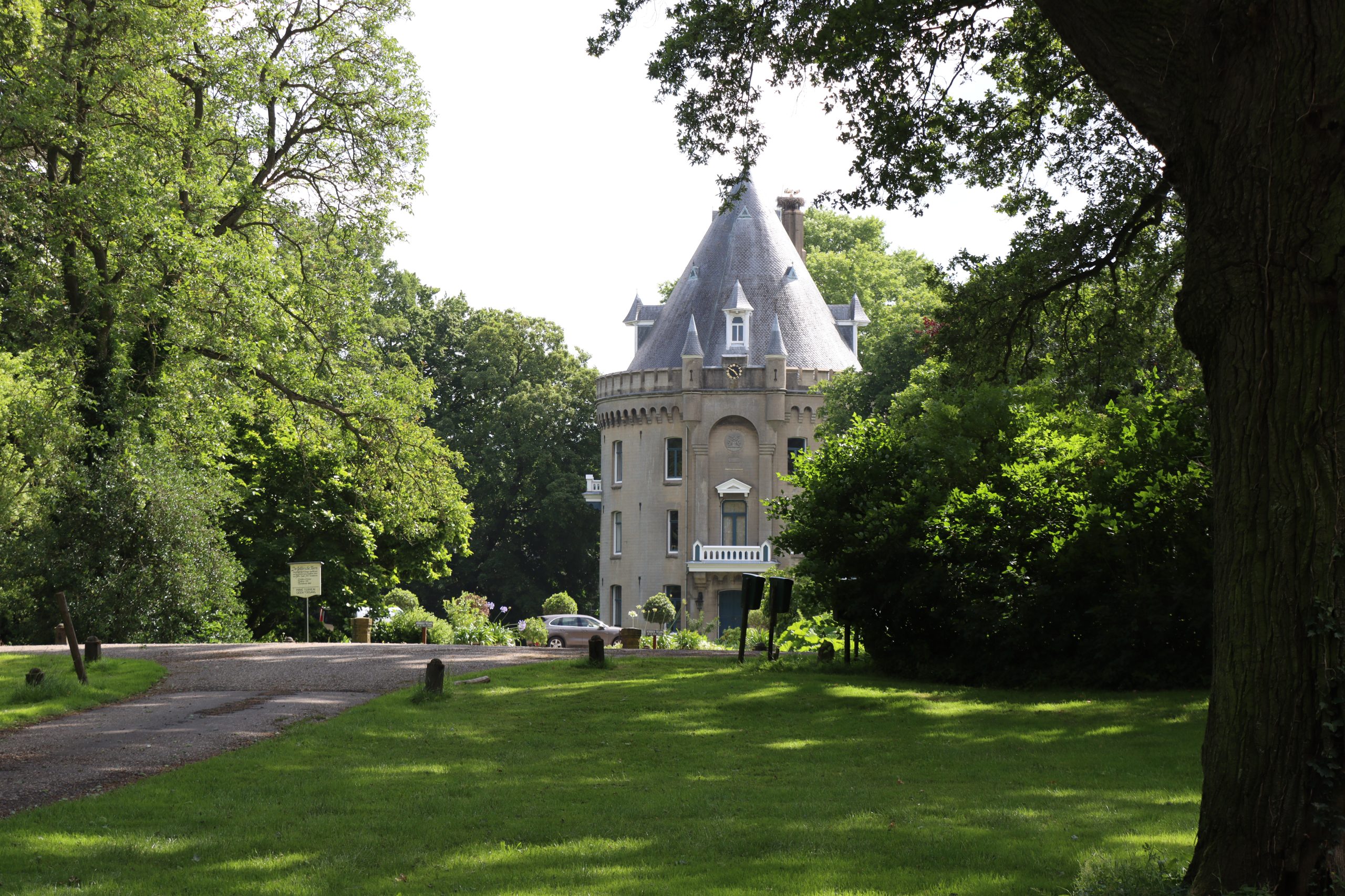 Kasteelachtig gebouw omringd door groen, met grasveld en grote bomen op een zonnige dag.