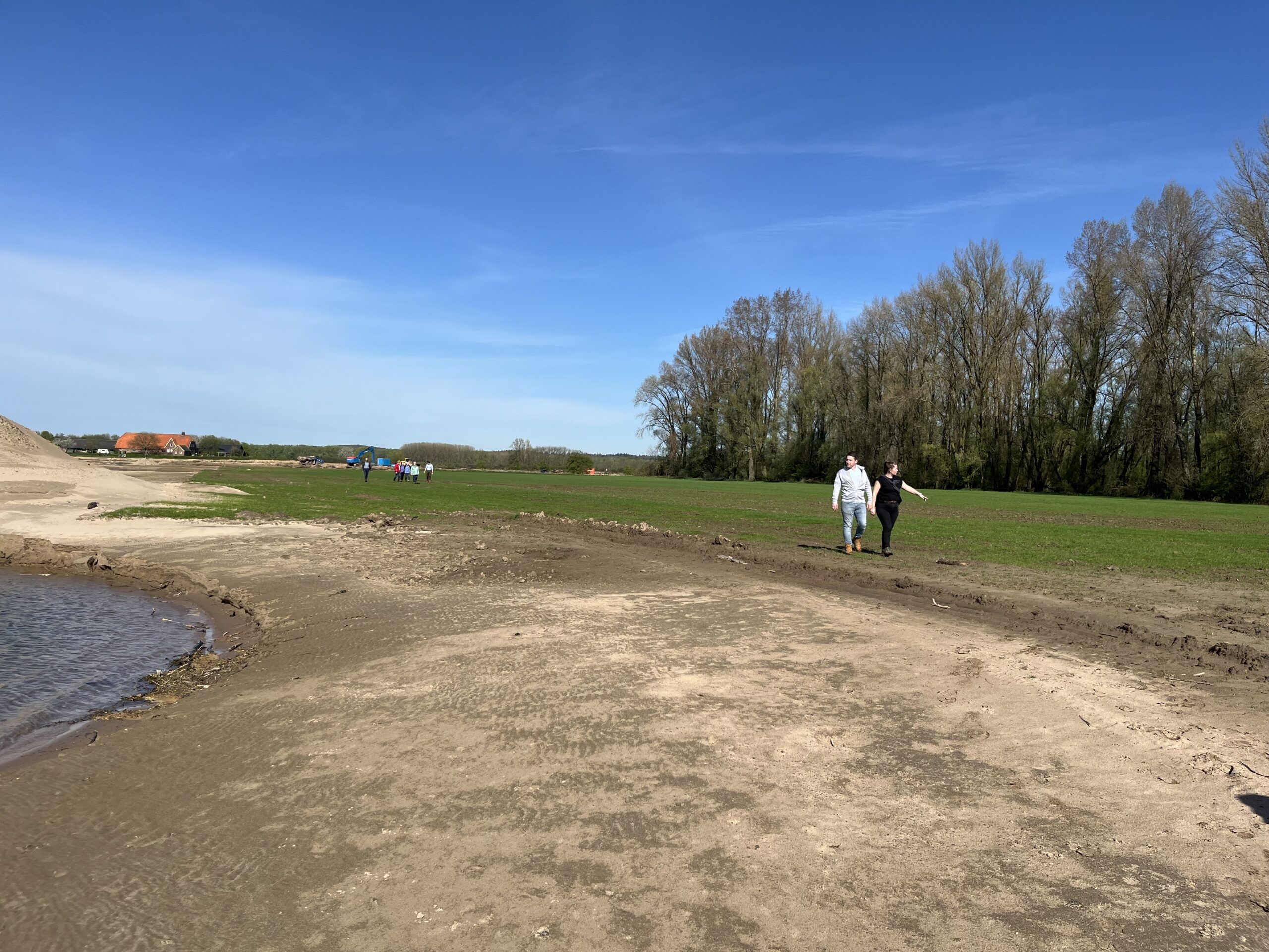 Een paar wandelt langs een modderige oever met bomen en velden op de achtergrond onder een blauwe lucht.
