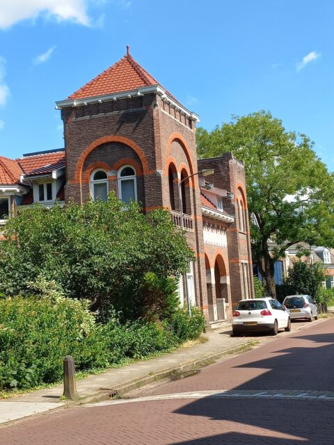 Rode bakstenen huis met toren en boogvensters, groene struiken en geparkeerde auto's naast straat.
