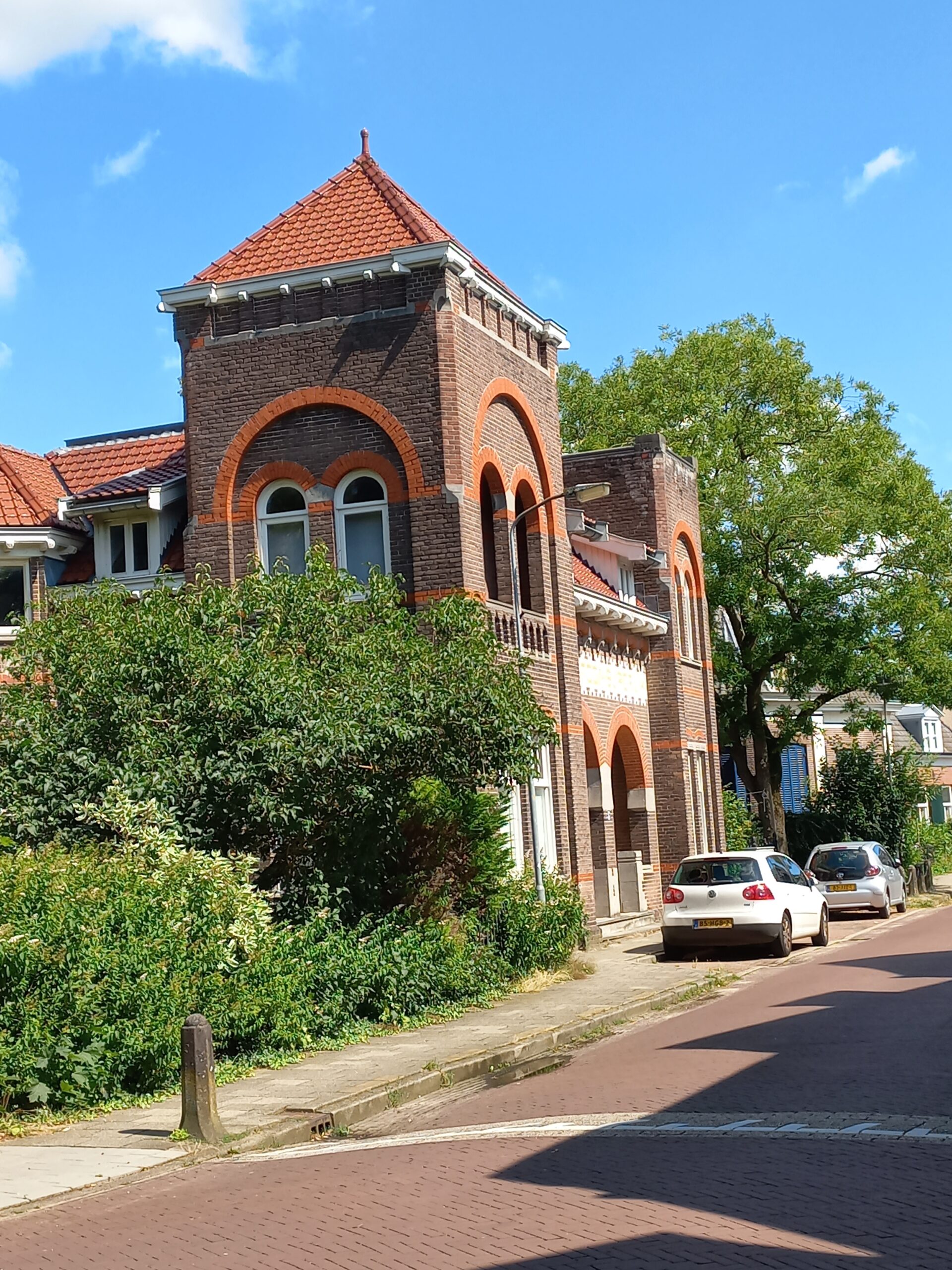 Rode bakstenen huis met toren en boogvensters, groene struiken en geparkeerde auto's naast straat.