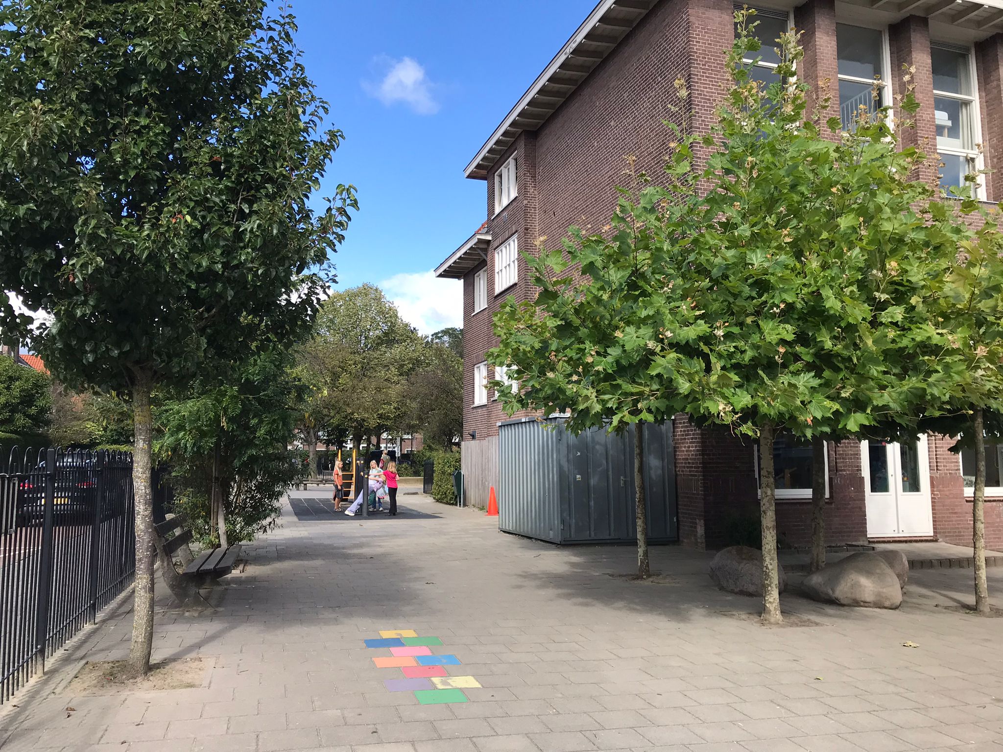 Schoolpleintje met bomen, hek, kleurrijke tegels en enkele kinderen spelend onder een blauwe hemel.