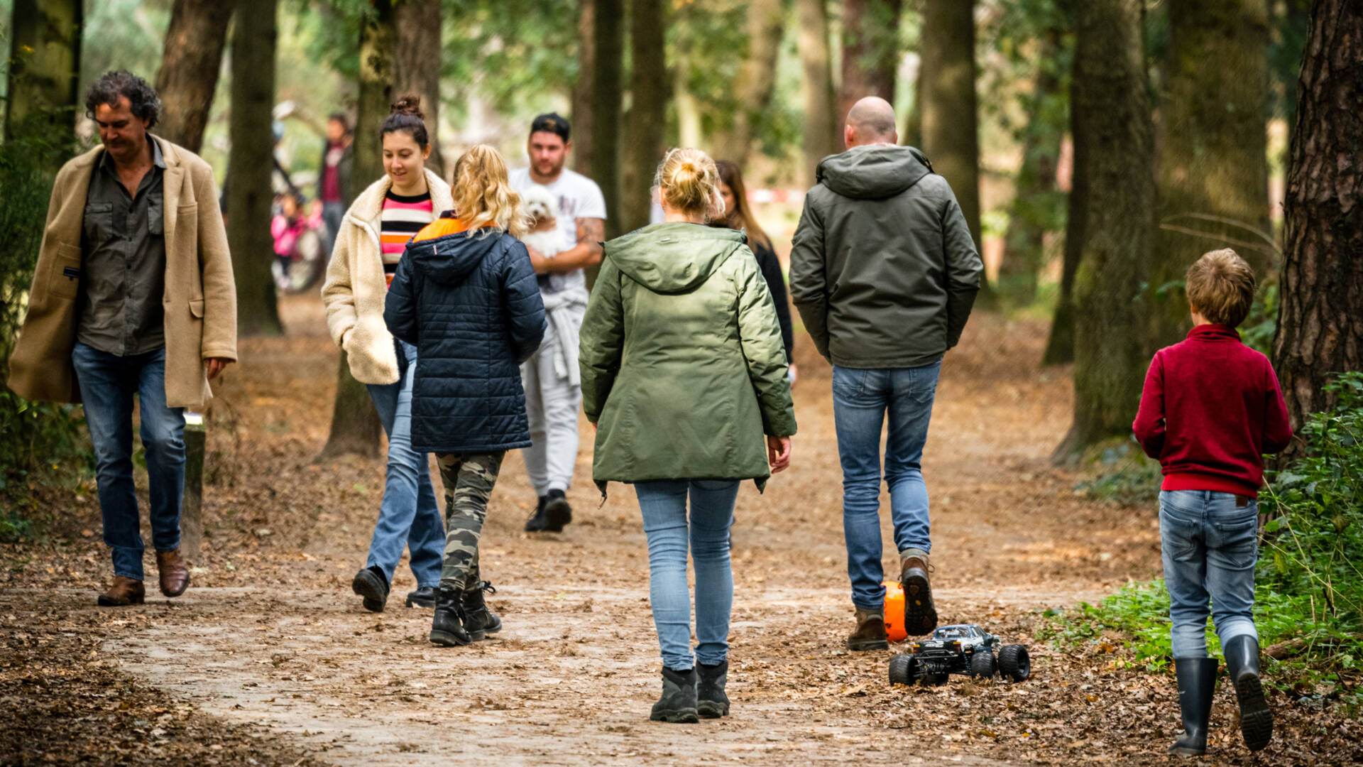 Mensen wandelen in een bos, een jongen speelt met een speelgoedauto op het pad.