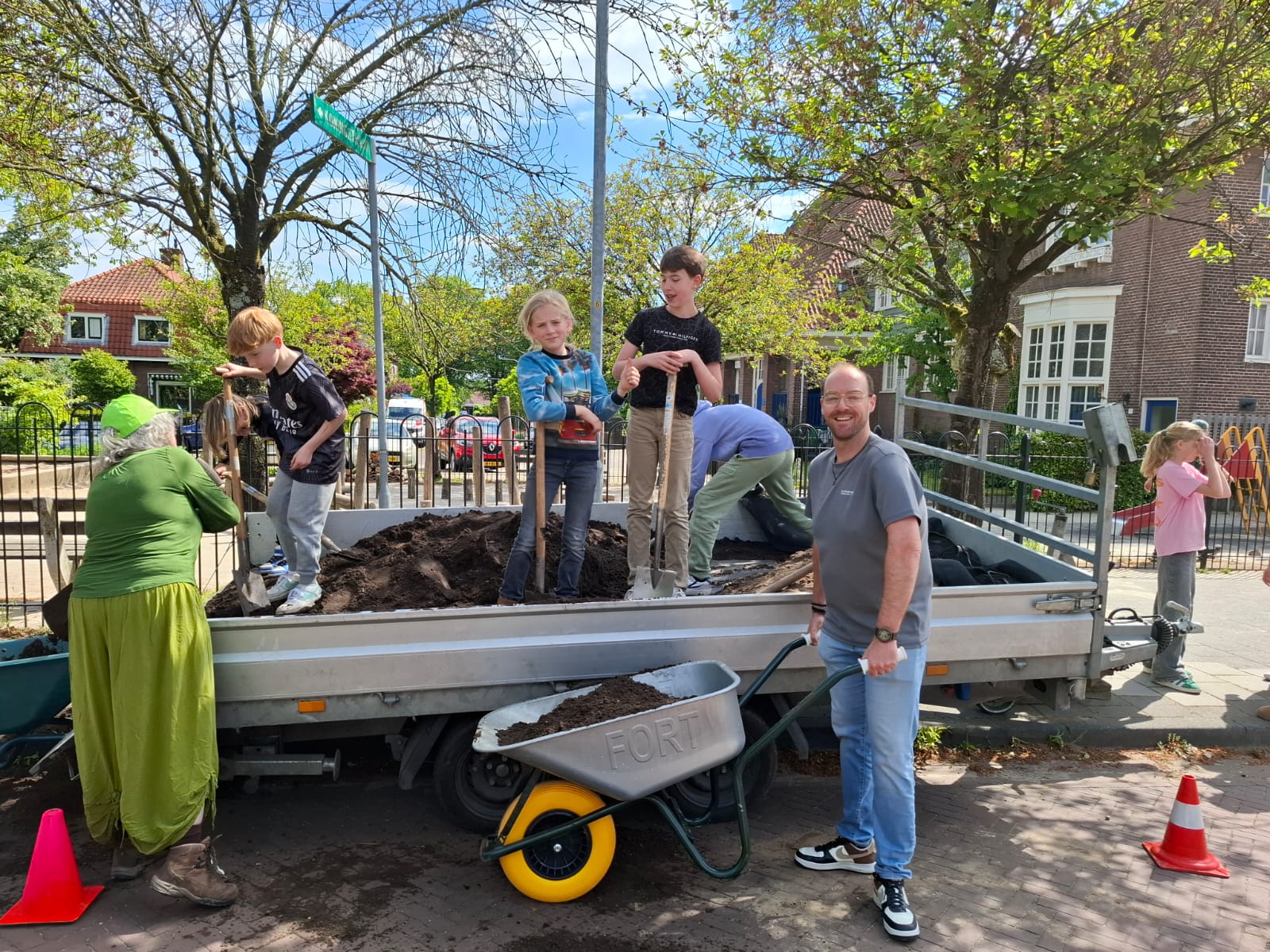 Kinderen en volwassenen scheppen aarde in een aanhanger op een zonnige dag in een woonwijk.