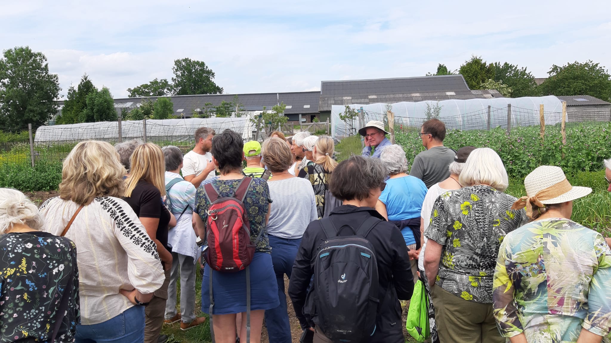 Groep mensen in buitenomgeving, luisterend naar een spreker naast serres en planten.