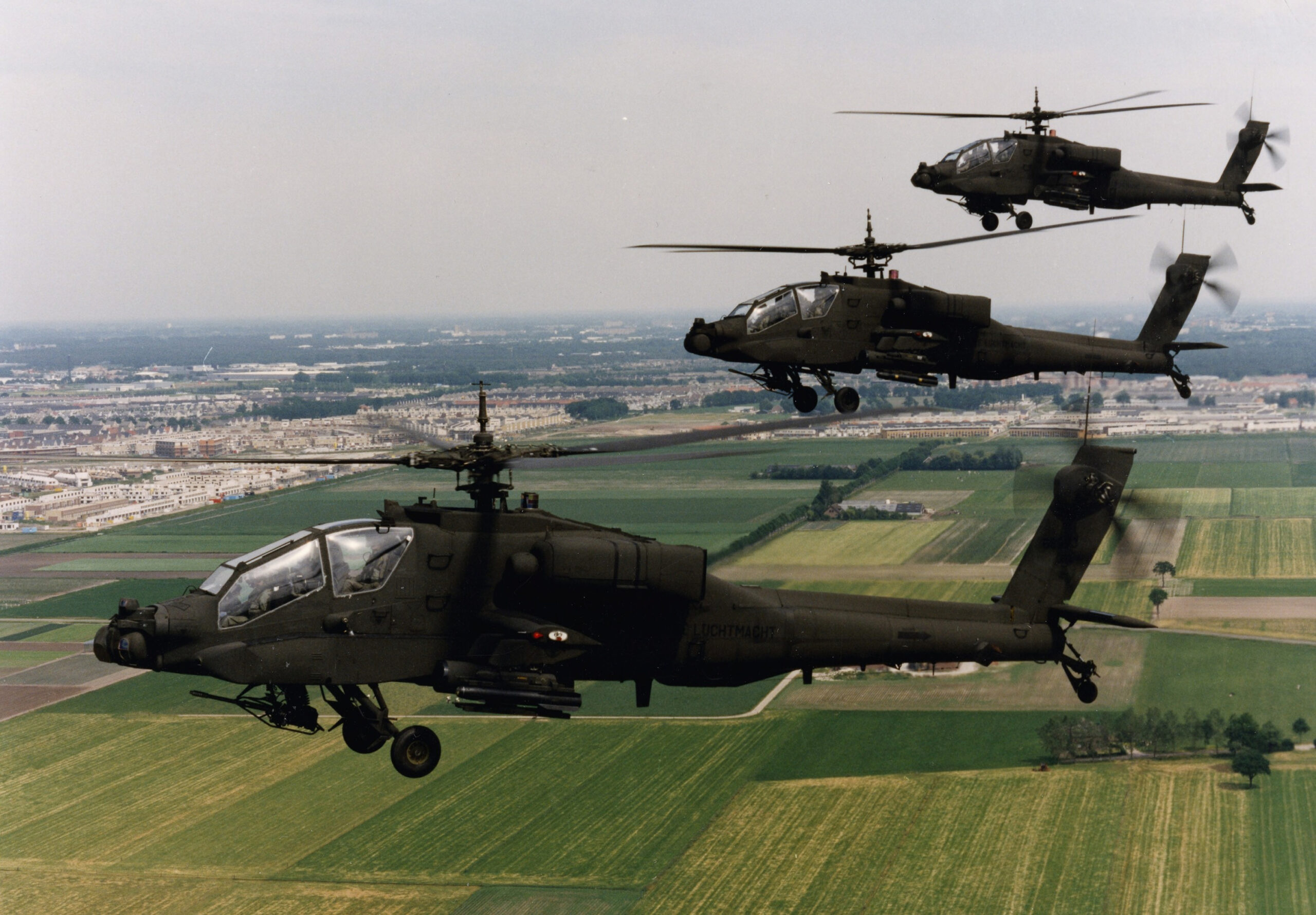 Drie Apache-helikopters vliegen over groene velden en een stad in de verte.
