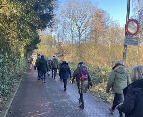 Groep mensen wandelt op een bospad; verkeersbord en bomen op de achtergrond.
