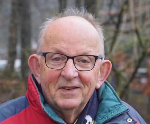 Een oudere man met bril en jas, buiten in een bosrijke omgeving.