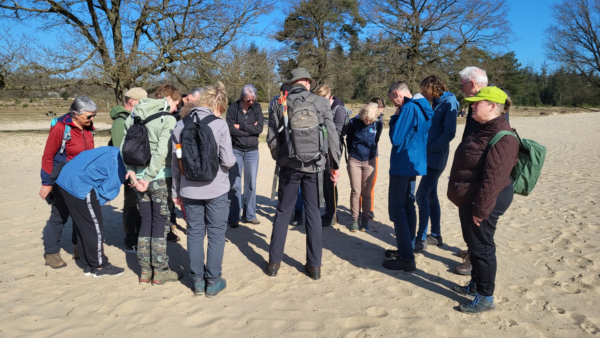 Groep mensen verzameld op een zandpad in een bos, in gesprek, met rugzakken en wandelschoenen.