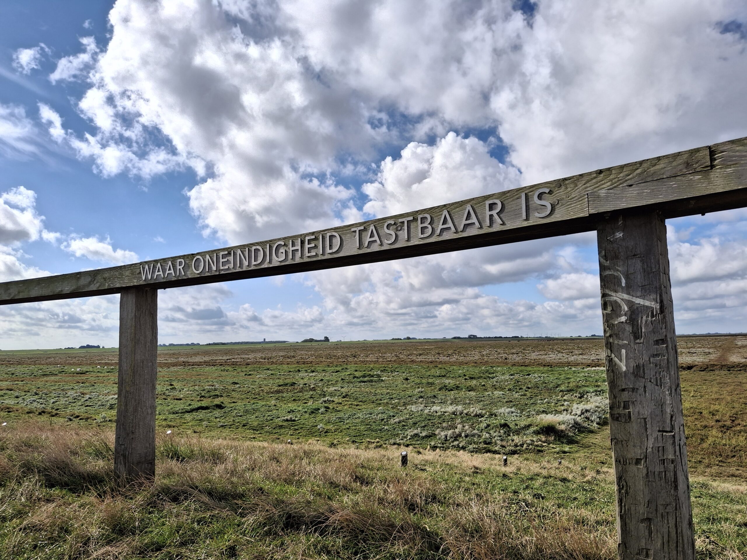 Houten poort met tekst "Waar Oneindigheid Tastbaar Is" overkijkt een groen landschap onder wolken.