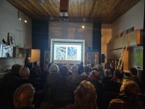 Een groep mensen kijkt naar een presentatie met dia's in een kleine zaal.