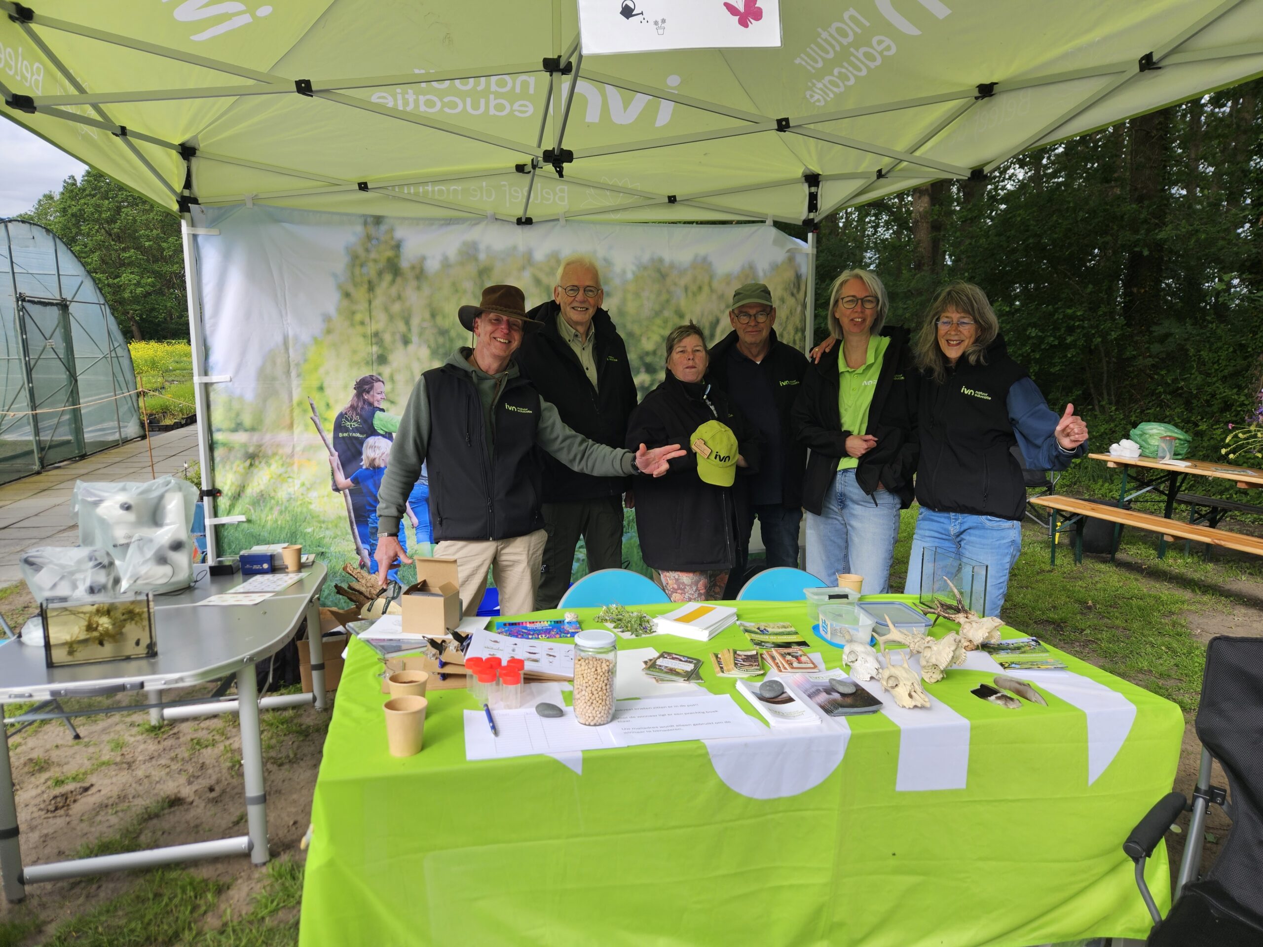 Groep mensen onder een partytent met natuurposters, omringd door educatieve materialen op een tafel.