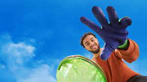 Man in oranje trui en paarse handschoen houdt groene vuilniszak onder een blauwe lucht vast.