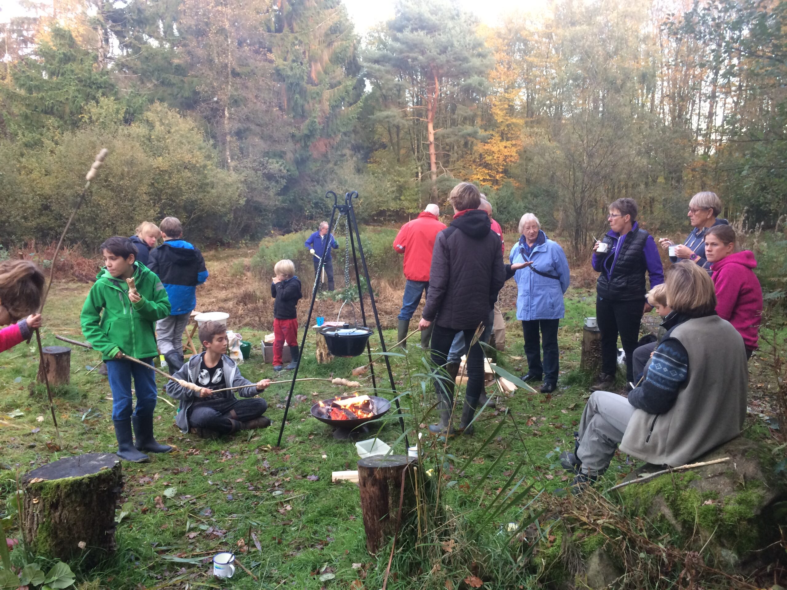 Mensen verzamelen rond een kampvuur in een bos; sommigen roosteren brood aan stokken.