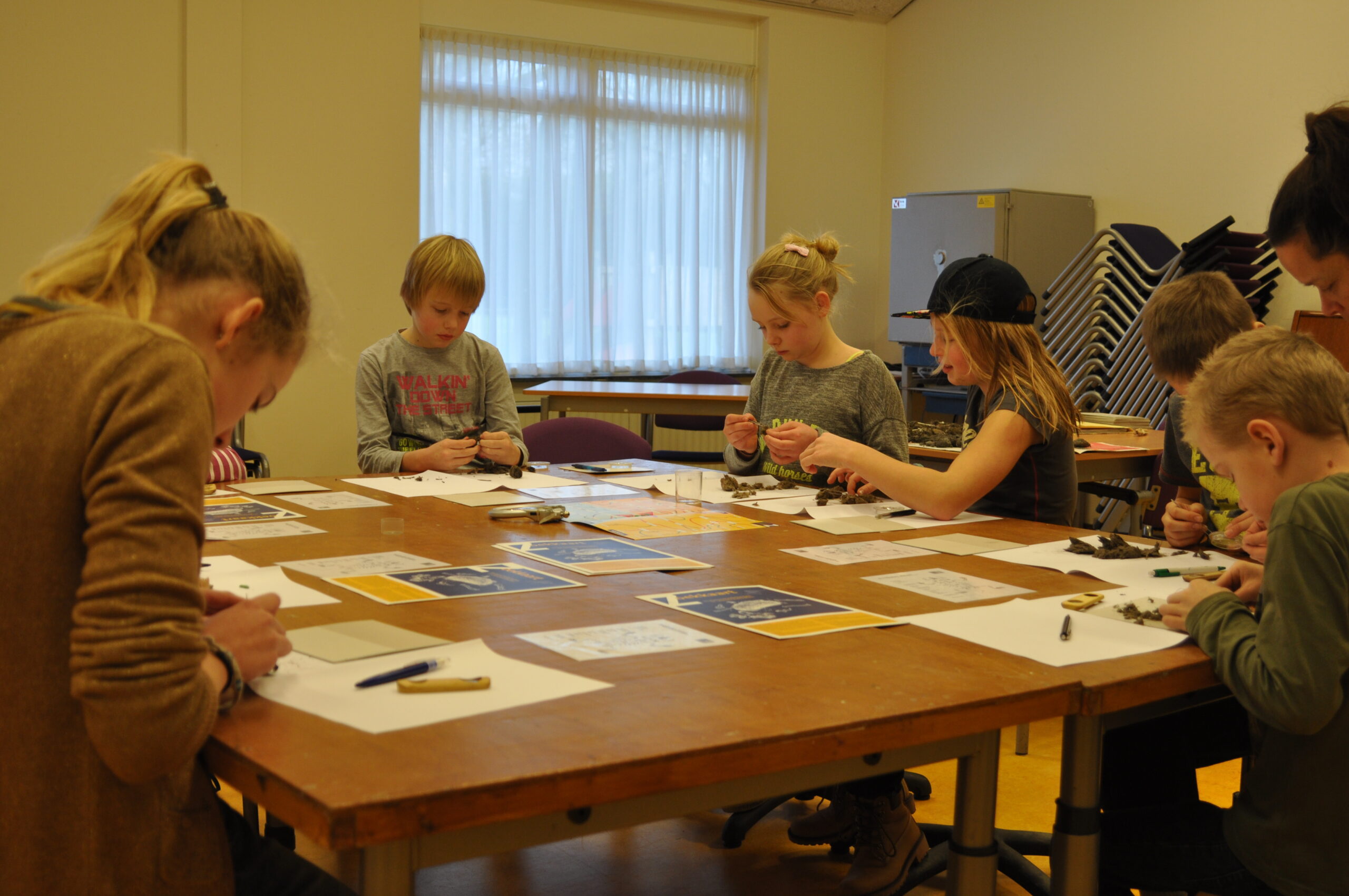 Kinderen knutselen aan tafel in klaslokaal, omringd door papieren en gereedschap.