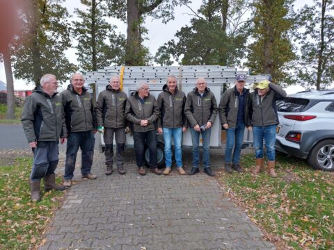 Groepje mannen in groene jacks poseren bij aanhangwagen met ladders, omringd door bomen en auto's.