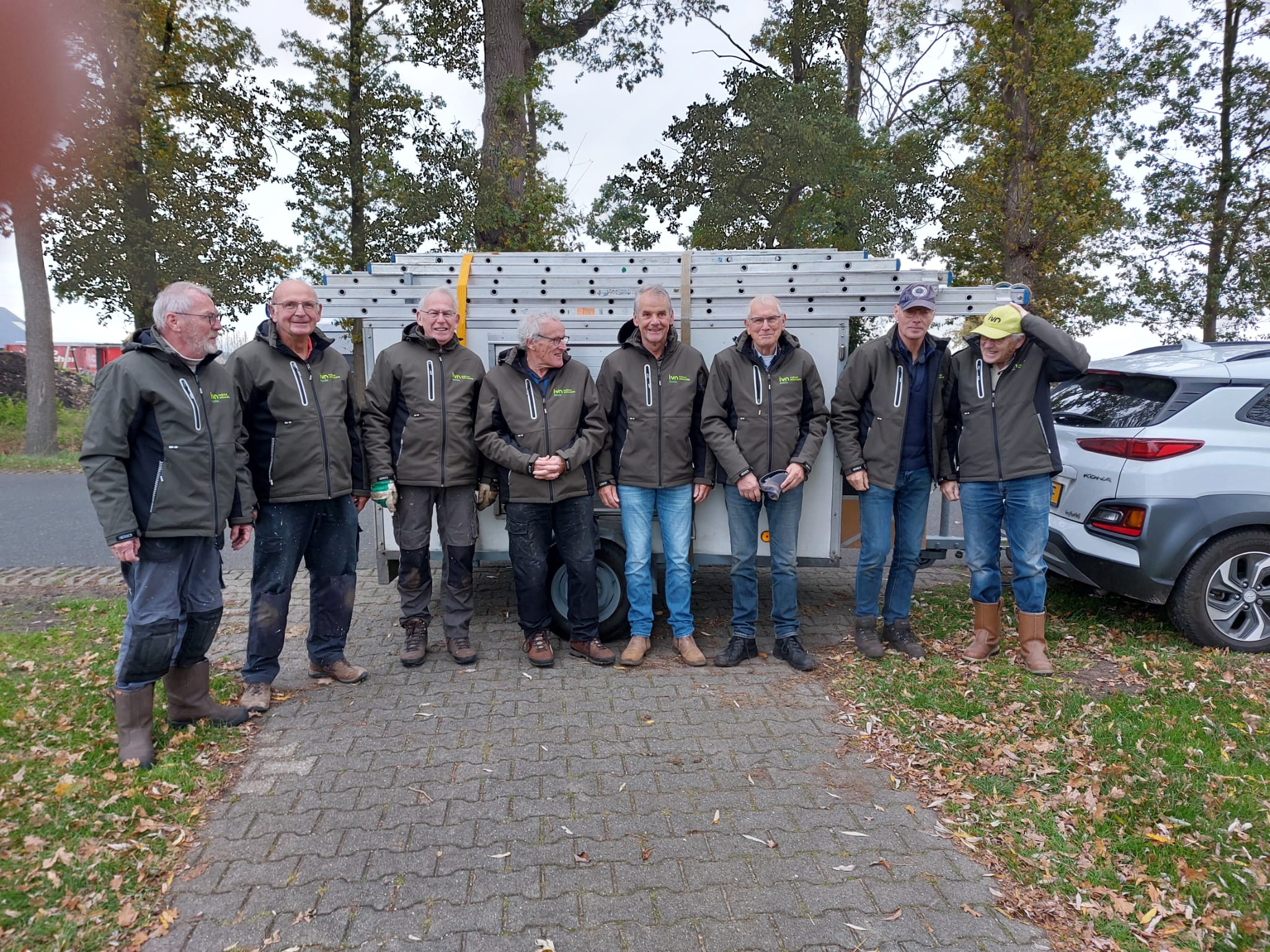 Groepje mannen in groene jacks poseren bij aanhangwagen met ladders, omringd door bomen en auto's.