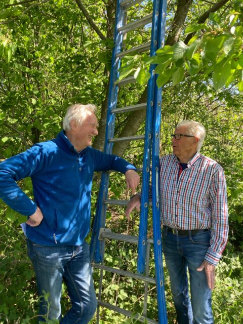 Twee mannen praten naast een ladder in een groene tuinomgeving.