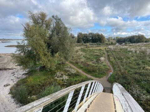 Brug leidt naar groen wandelpad langs rivieroever met bomen en wegwijzers onder bewolkte lucht.