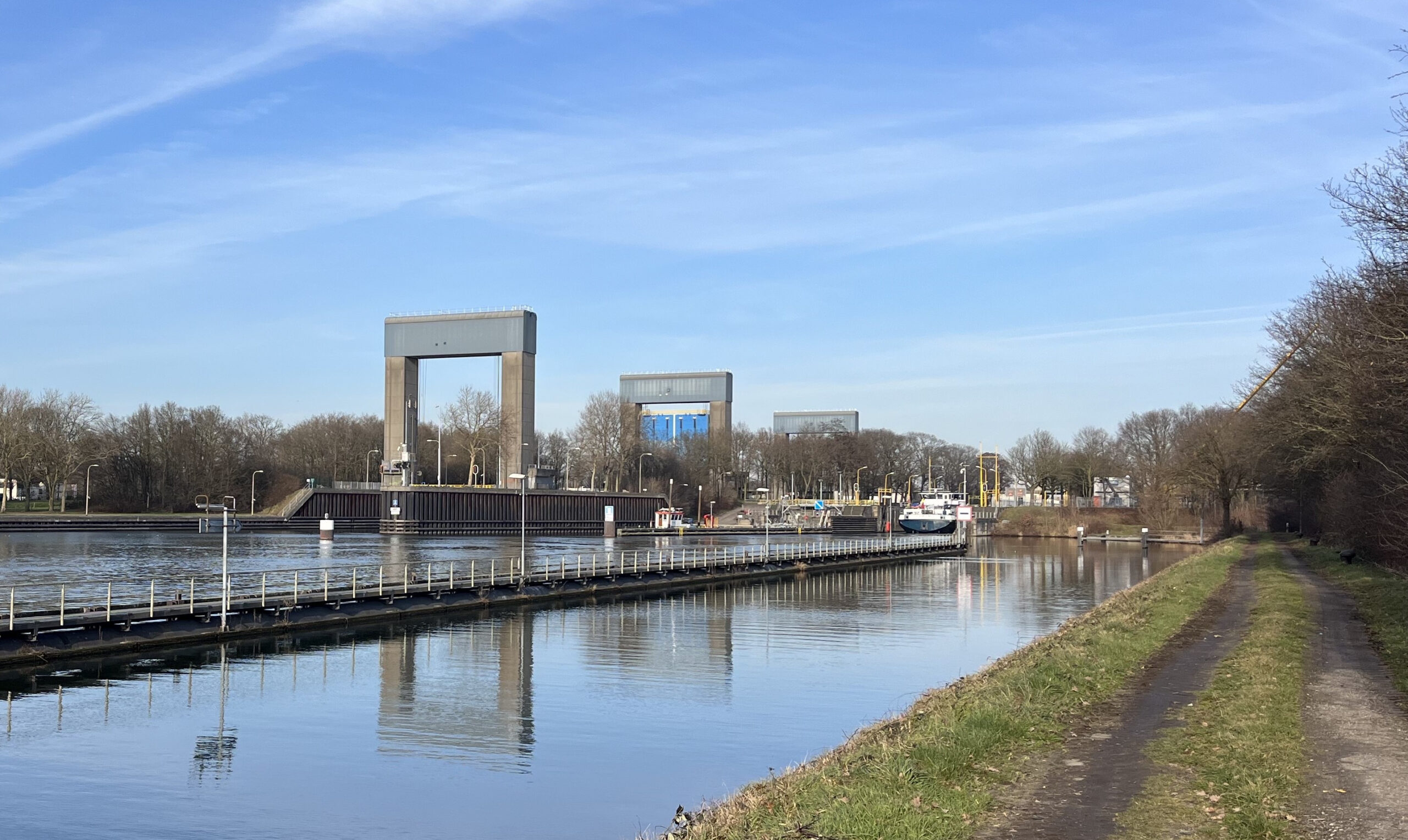 Kanaal met sluizen en een boot, geflankeerd door een pad en bomen onder een blauwe hemel.