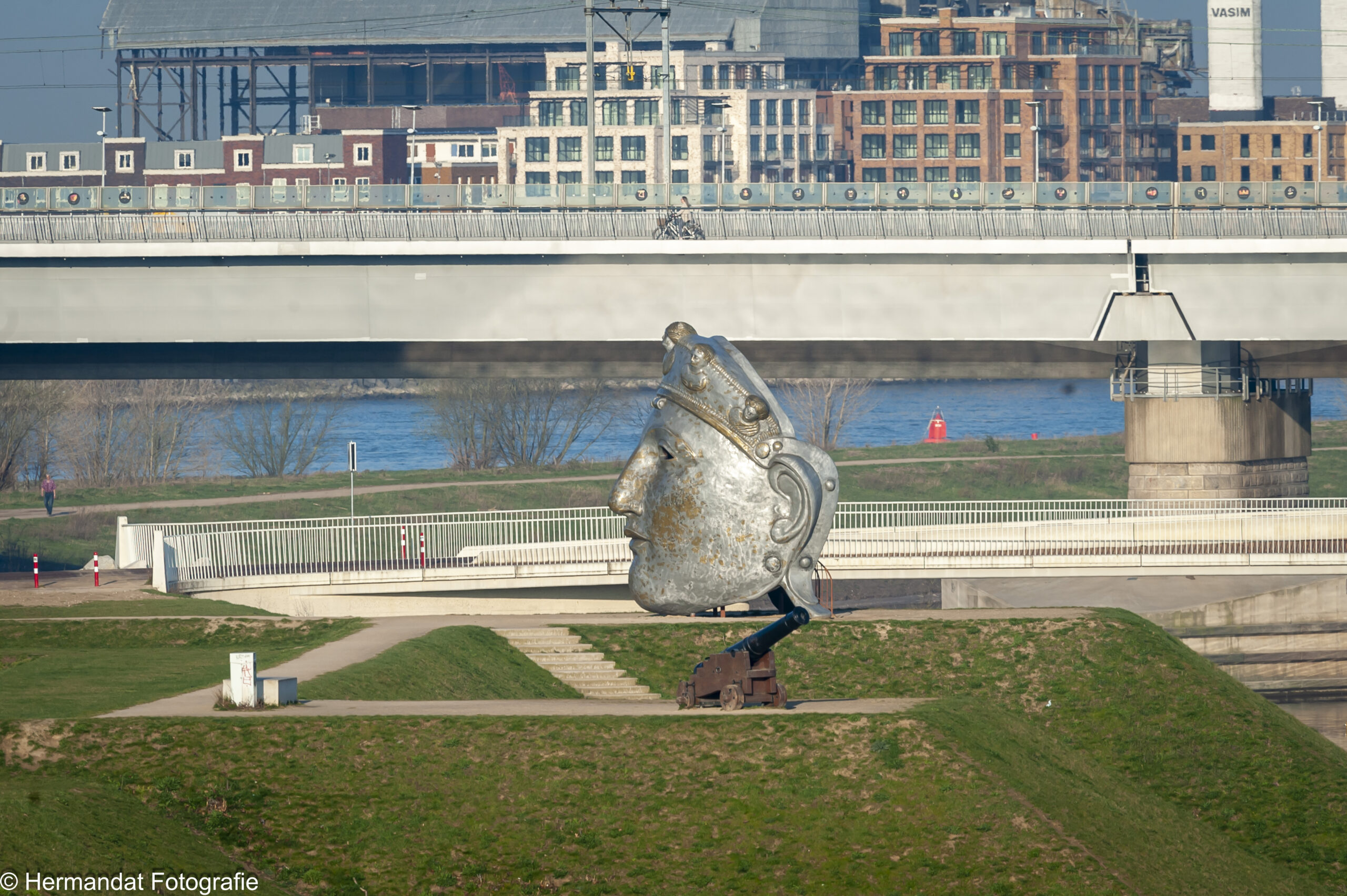 Groot standbeeld van een Romeinse helm naast een kanon, voor een moderne brug en stadsgebouw.