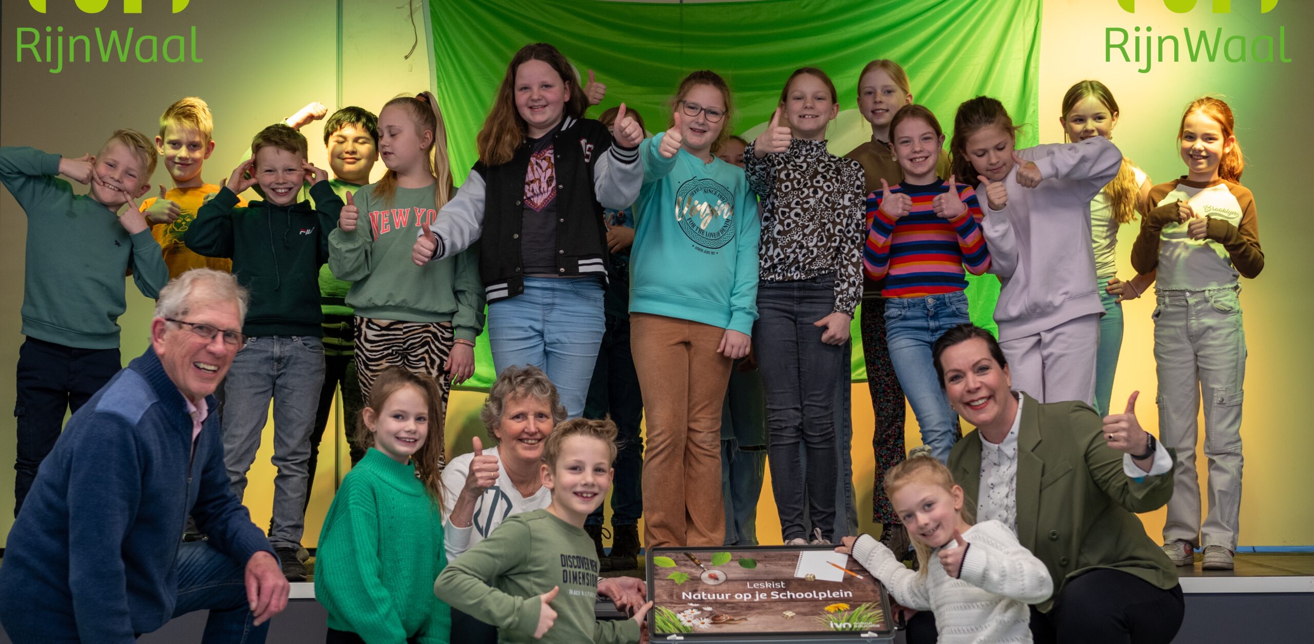Groep kinderen en volwassenen glimlacht en toont duimen omhoog bij "Natuur op je Schoolplein" poster.