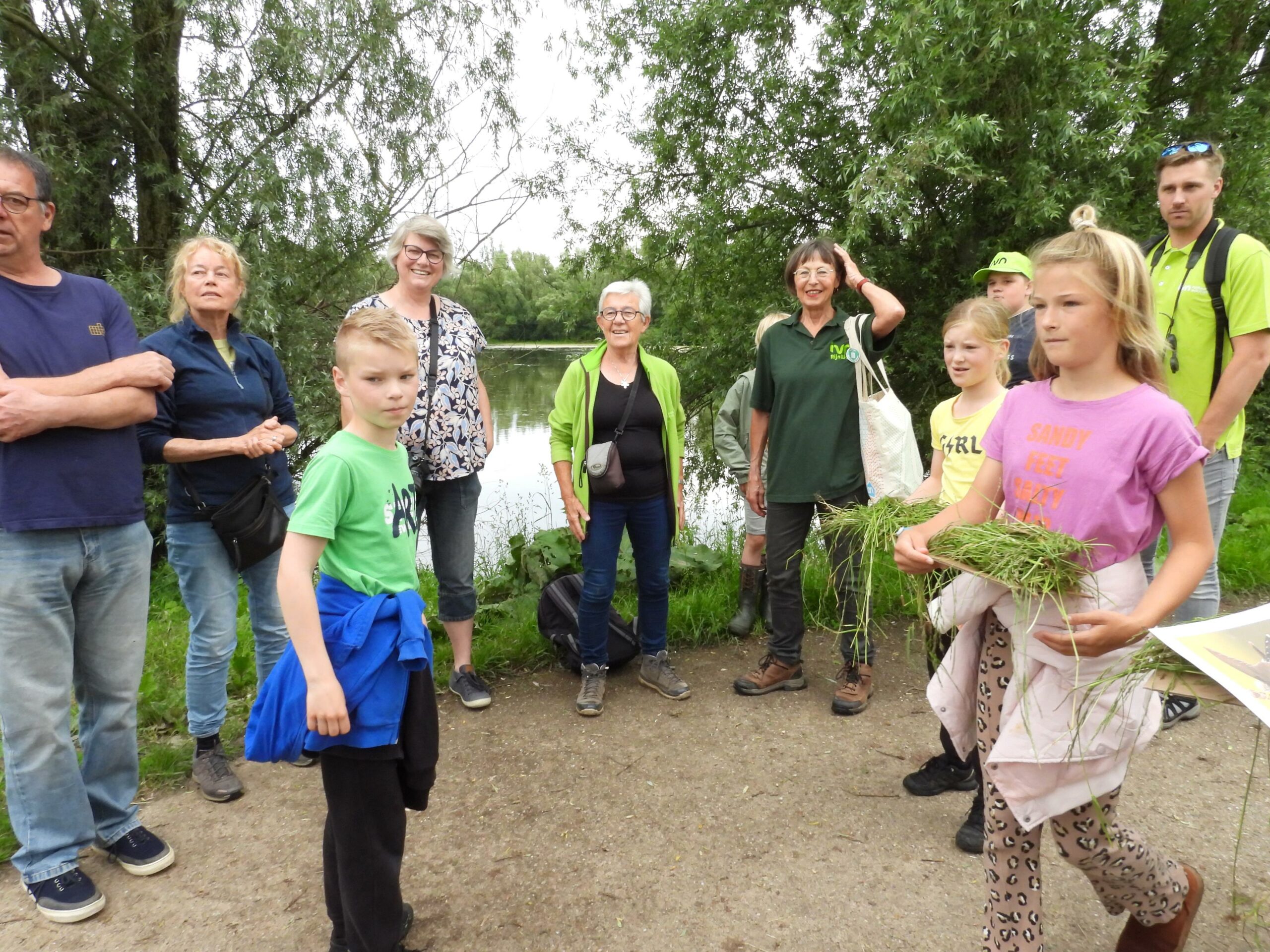 Een groep mensen, waaronder kinderen met gras, staat buiten bij een vijver en bomen.