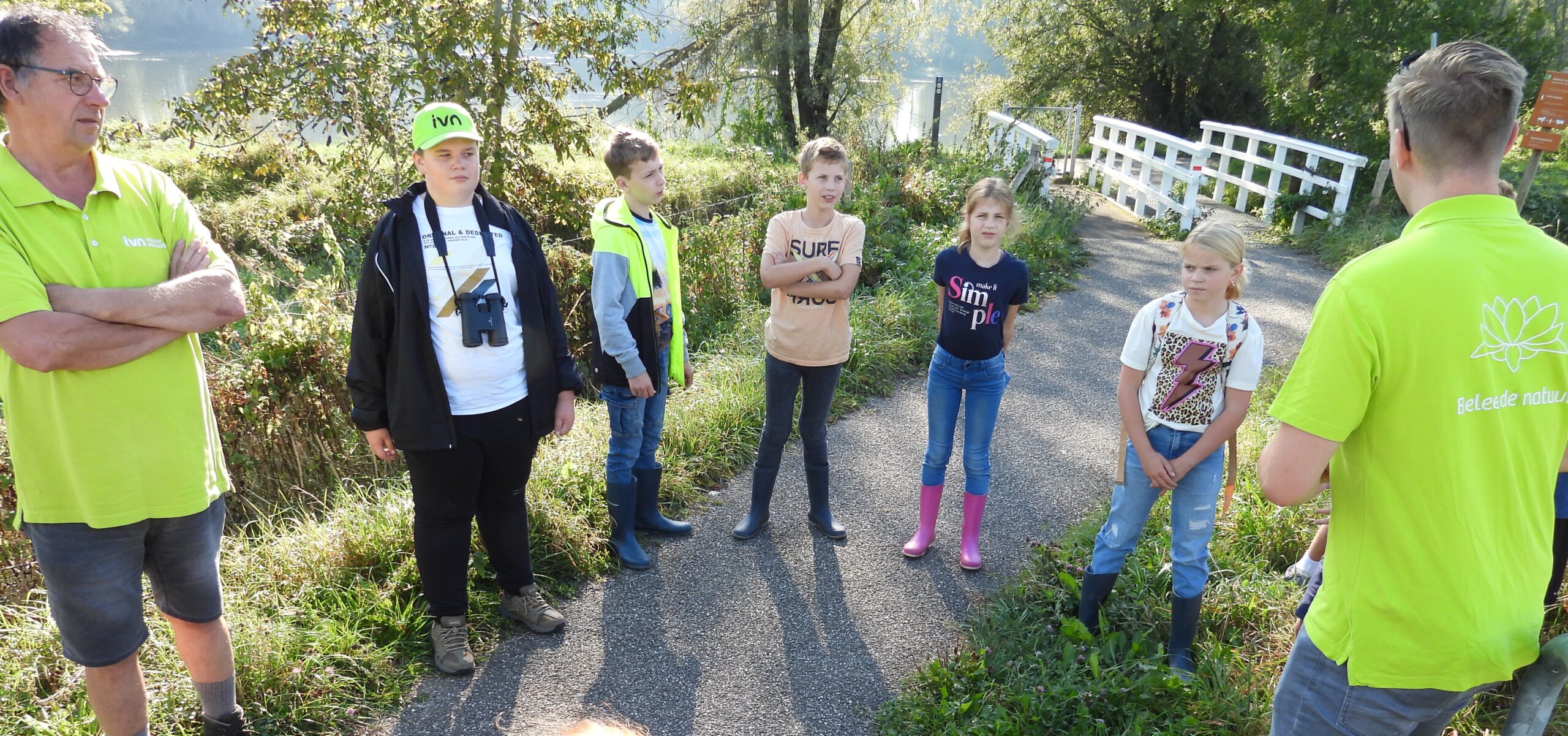 Groep kinderen luistert naar gids in groene shirt op natuurpad met een brug op de achtergrond.