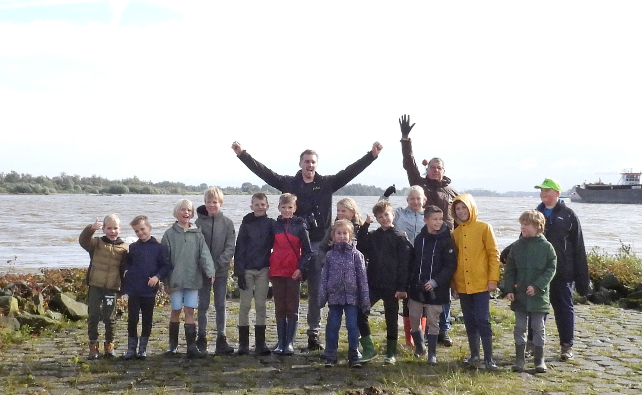 Groep mensen in regenjassen en laarzen poseert vrolijk bij een rivier met een schip op de achtergrond.